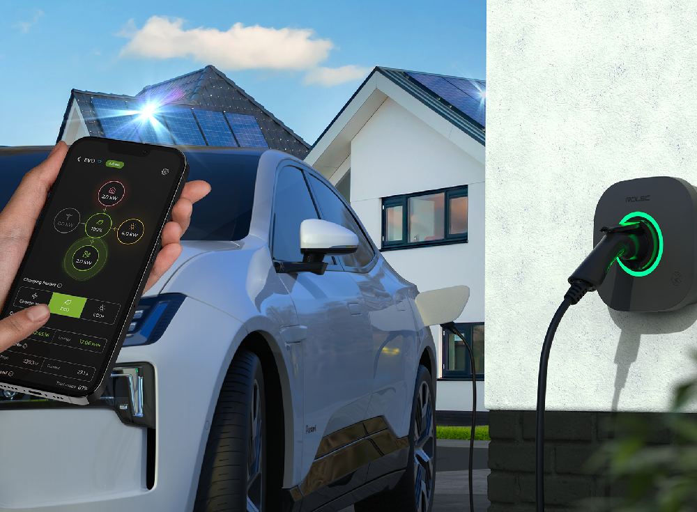 Electric car charging via a wall-mounted Rolec EVO EV charger, with a smartphone displaying the charger’s control app and solar panels on the house roof
